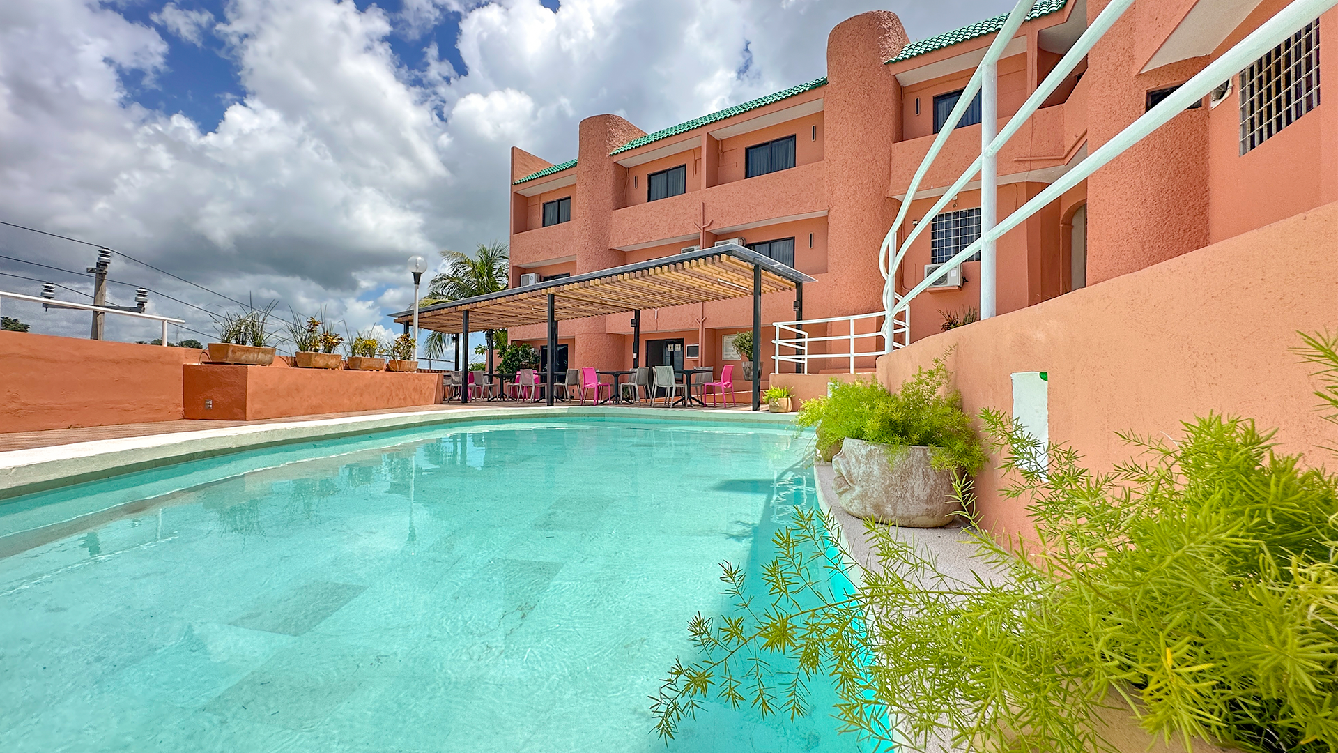 A turquoise swimming pool in front of a multi-story pink hotel building with a wooden pergola under a cloudy sky.