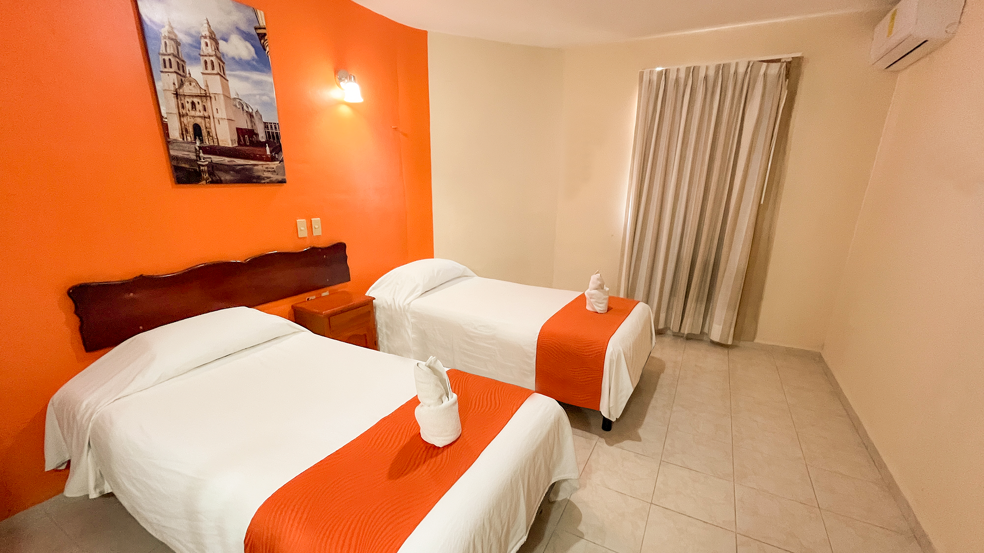 Hotel room with two twin beds, white linens with orange runners, and an orange accent wall with a wall art piece.