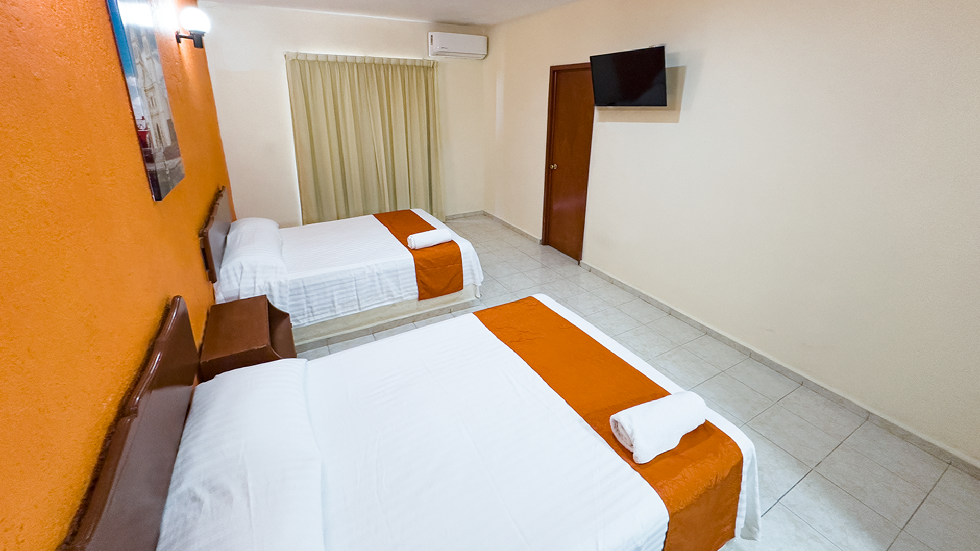 A hotel room with two white beds, orange accent wall and bed runners, tiled floors, a wall-mounted TV, and a window.