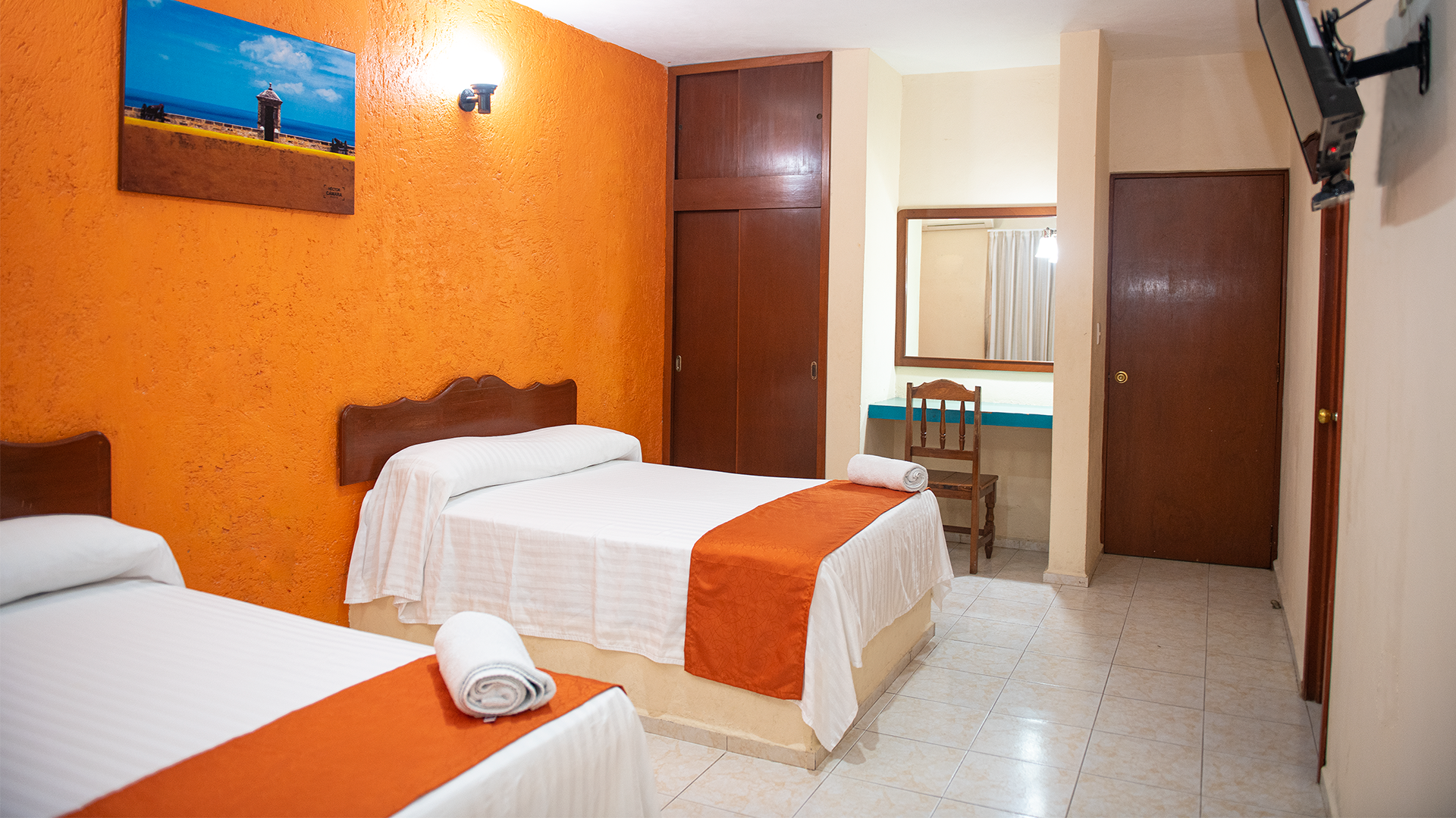 A hotel room with two beds, orange walls, tiled floors, a wooden closet, and a television mounted on the wall.