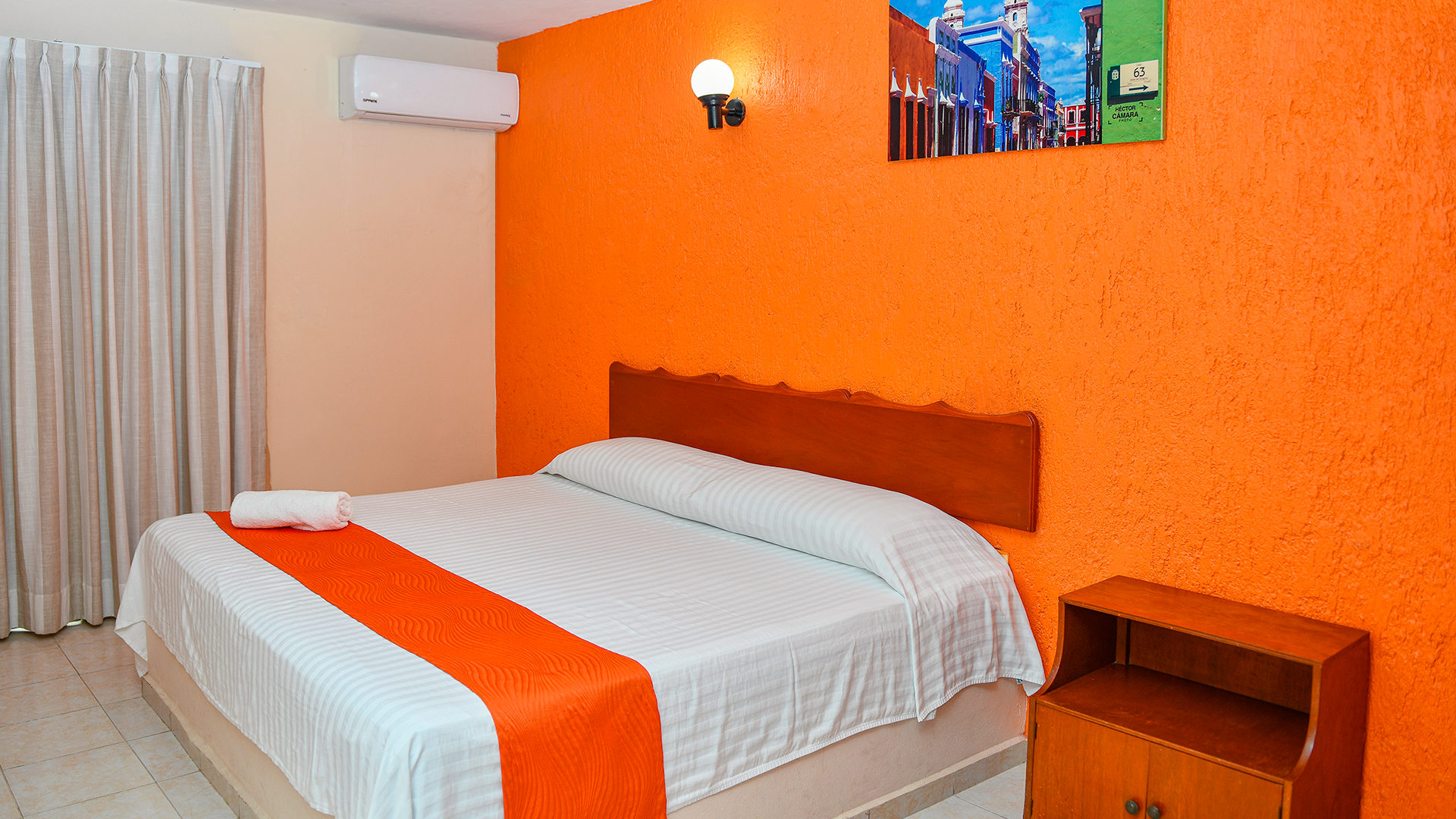 A bedroom with a bed featuring an orange runner, a vibrant orange accent wall, a wooden nightstand, and window curtains.