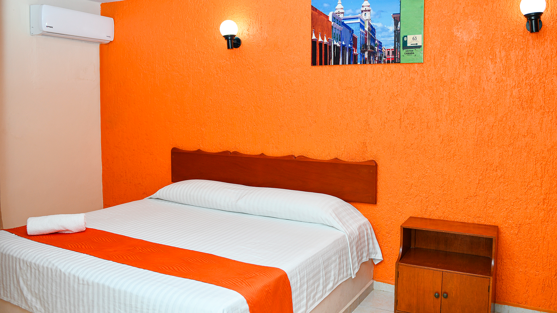 A bedroom with an orange textured wall, a white bed with an orange runner, a wooden nightstand, and two wall lamps.