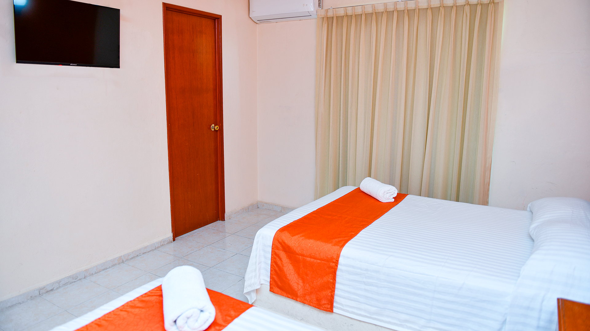 A hotel room with two white beds featuring orange runners, a wooden door, a wall-mounted TV, and light curtains.