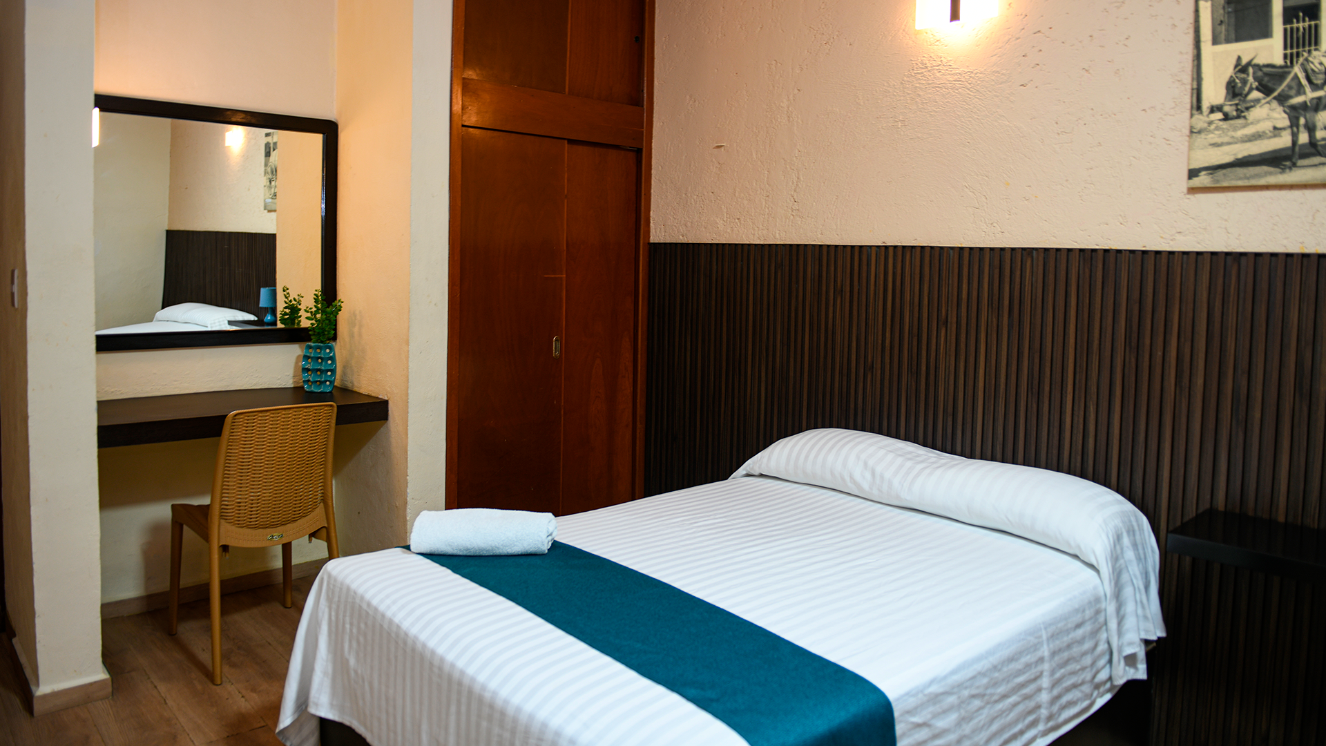 A hotel room with a neatly made bed, a small desk with a chair, a wall mirror, and a wooden closet against a dark wall.