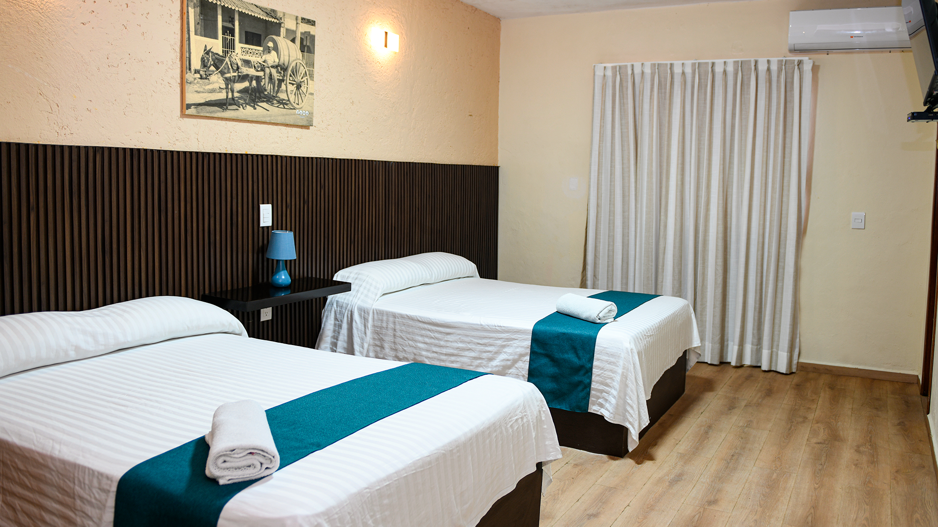 A hotel room with two beds featuring white bedding and teal runners, dark wood headboards, and a light-colored wood floor.