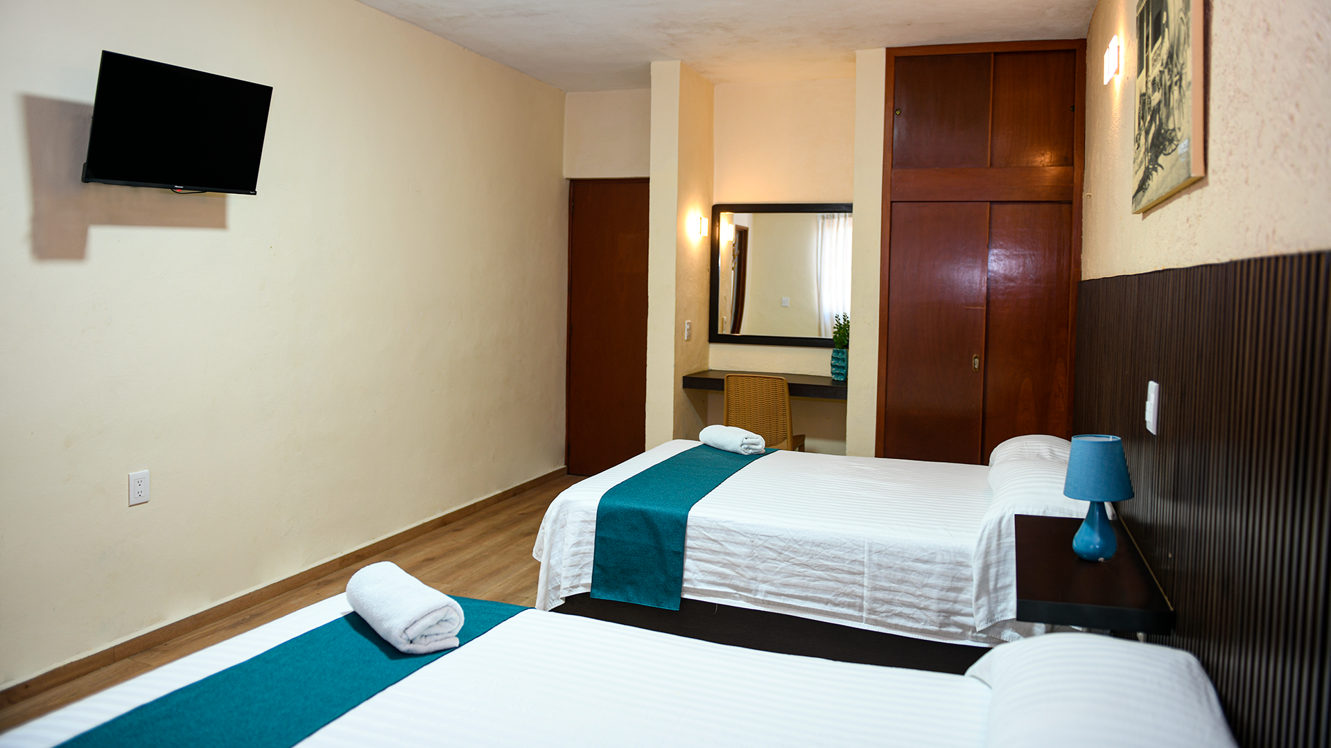 A hotel room with two beds featuring white linens and teal runners, a wall-mounted TV, and a wooden wardrobe.