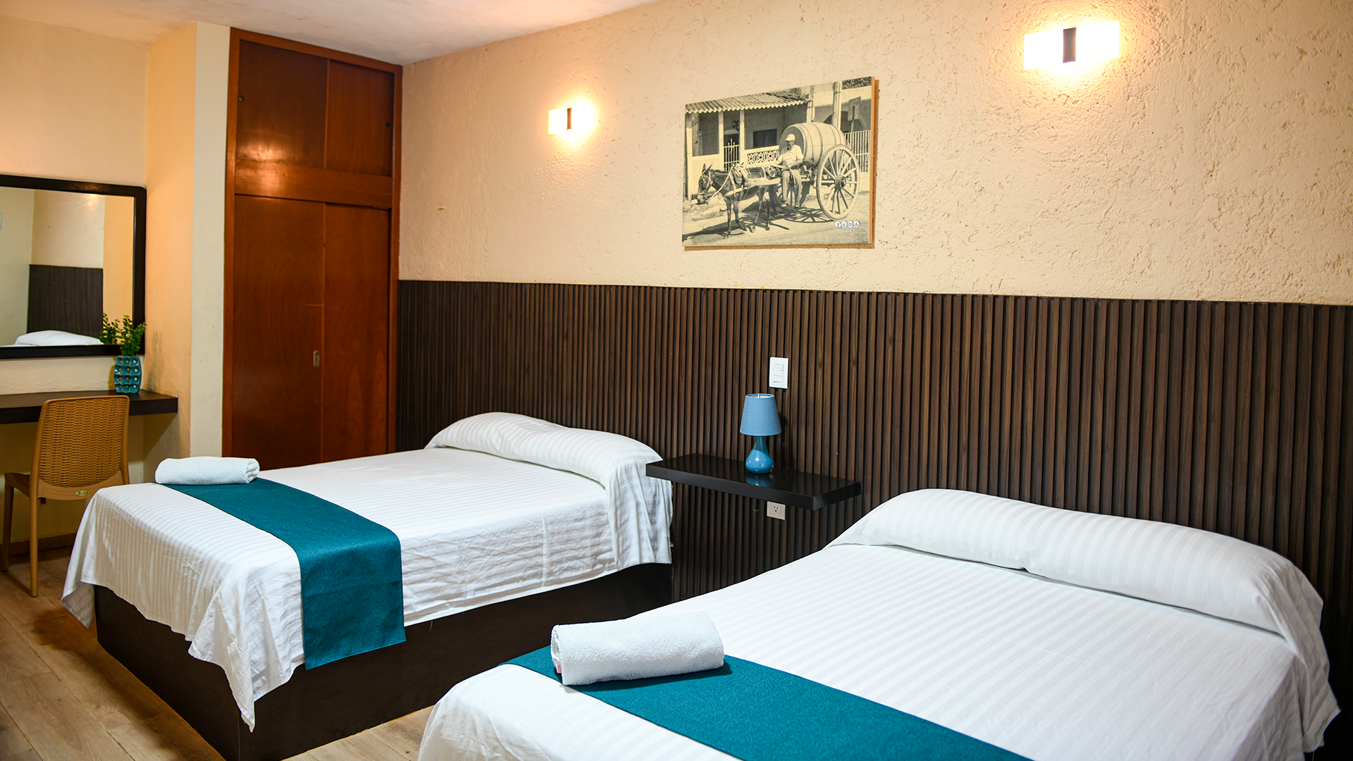 A hotel room featuring two beds with white linens and teal runners, a wooden wardrobe, and a desk against a textured wall.