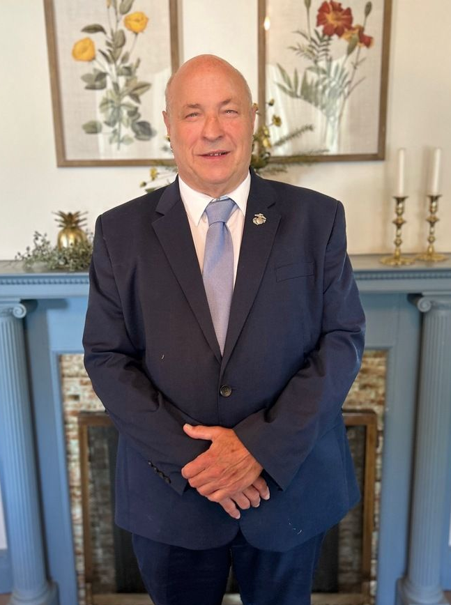 Man in blue suit, arms crossed, standing in front of fireplace, smiling. Flowers and candles in the background.