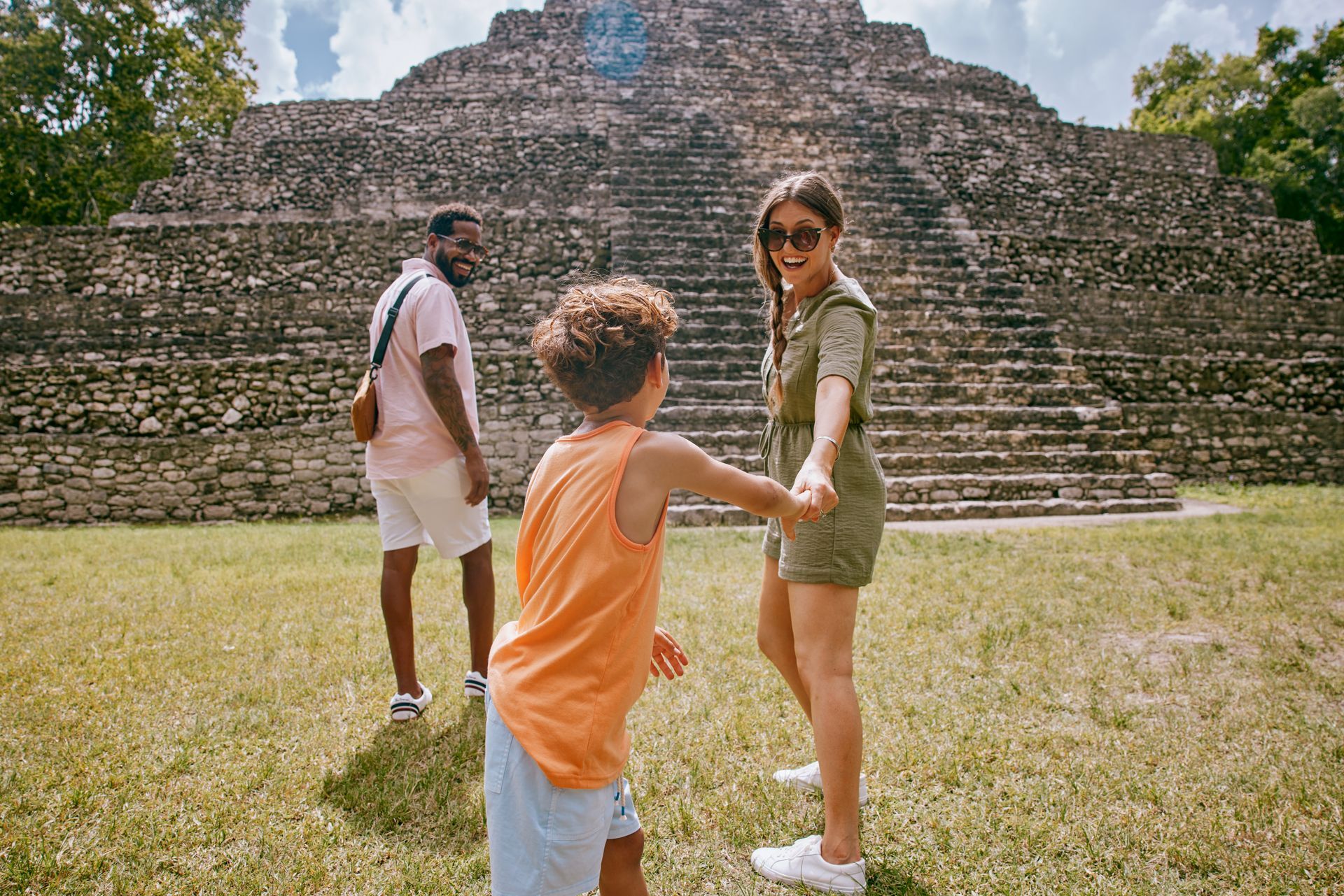 Three tourists standing before a stepped stone pyramid in a grassy ruin site