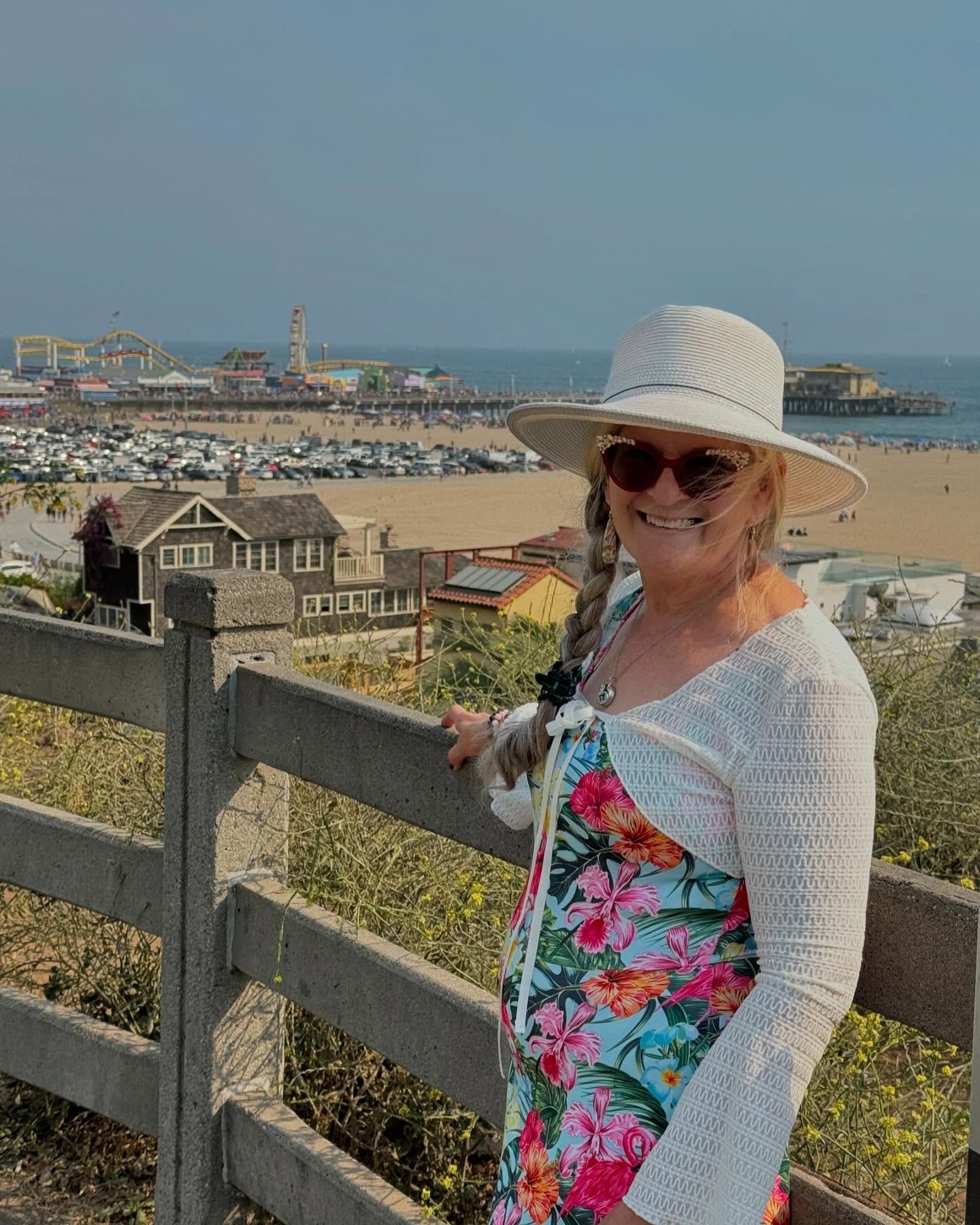 Woman in floral dress and sunhat smiling by a fence overlooking a beach town and harbor.