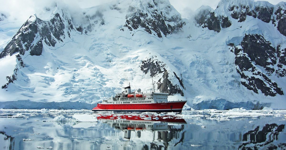 Red cruise ship on calm icy water below snowy mountain cliffs and their reflection