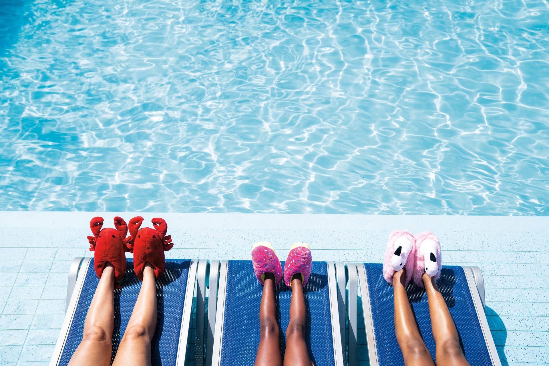 Four pairs of feet in colorful socks rest on lounge chairs beside a bright swimming pool.