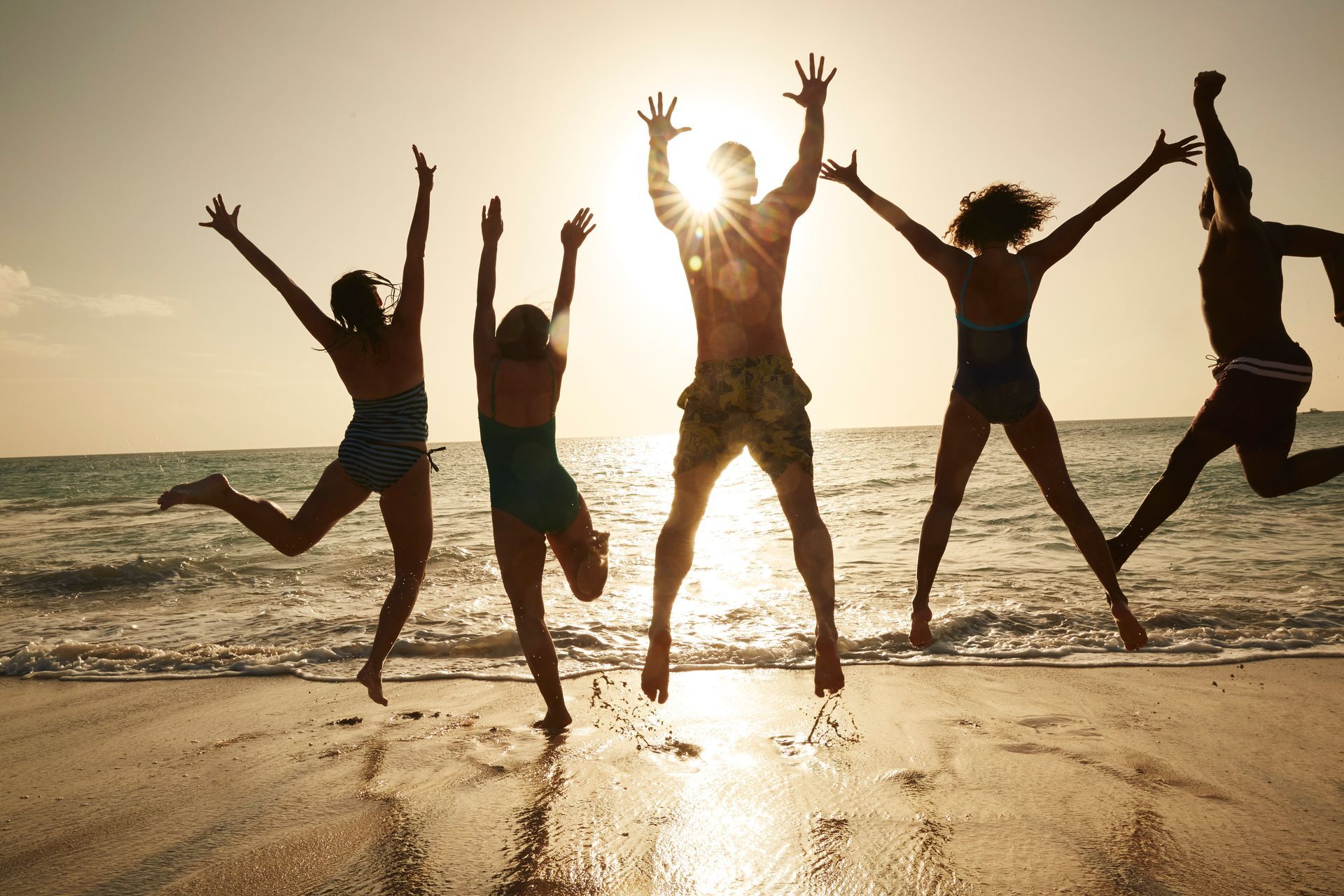 Five friends jumping with arms raised on a beach at sunset