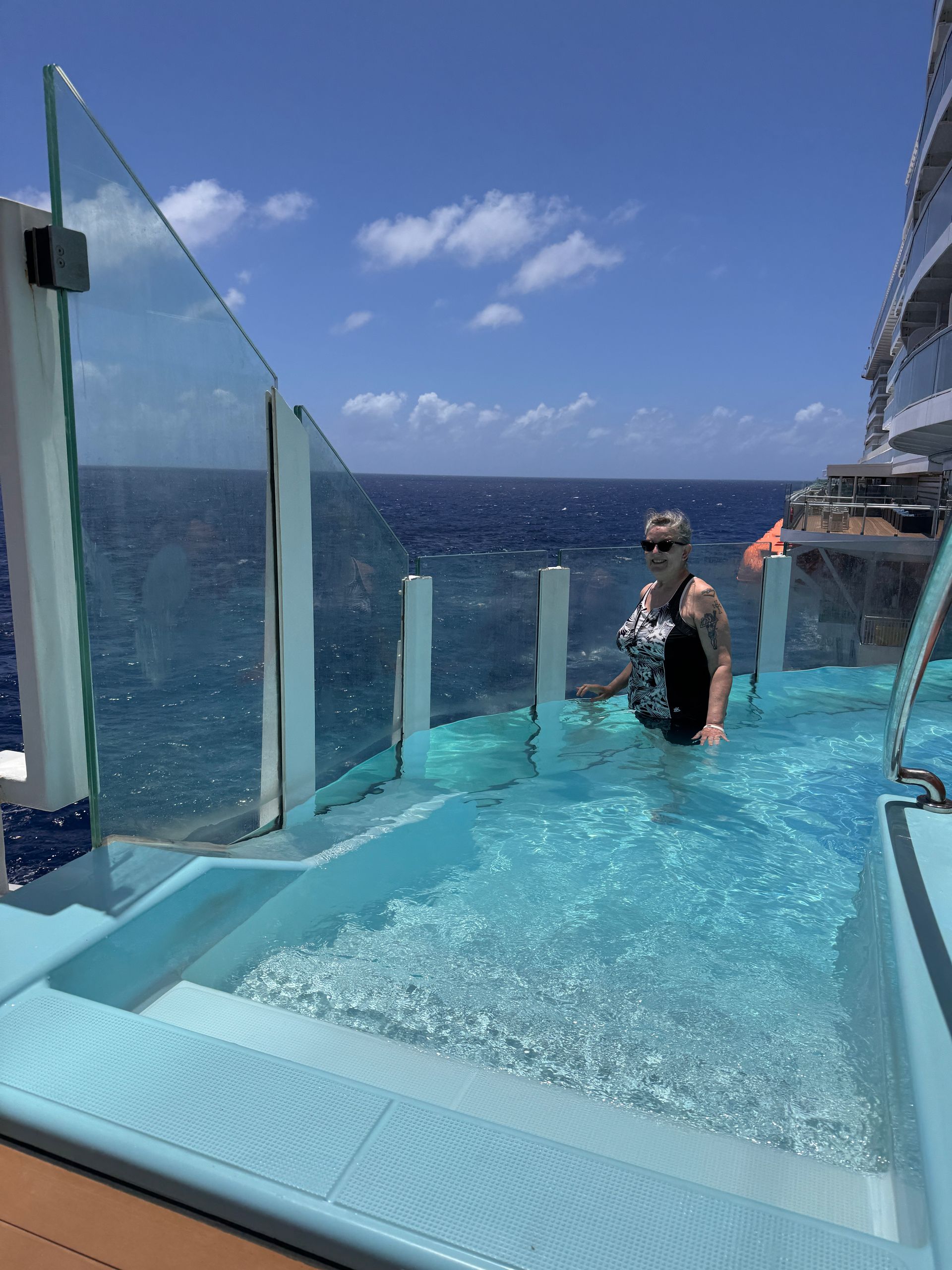 Person standing in a clear-water infinity pool beside glass panels overlooking the ocean and blue sky