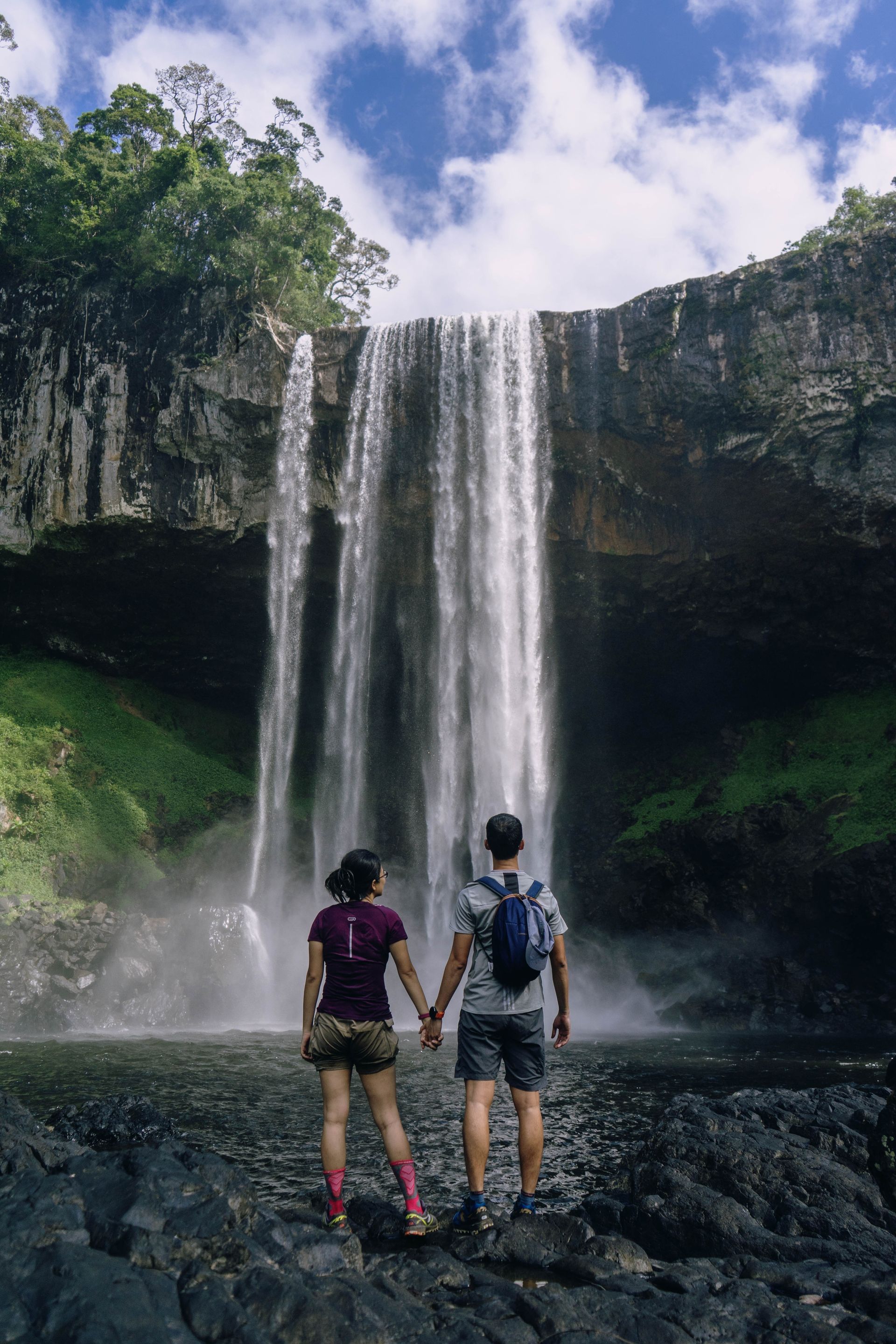 Two hikers stand on rocks before a tall waterfall and cliff under a cloudy sky.