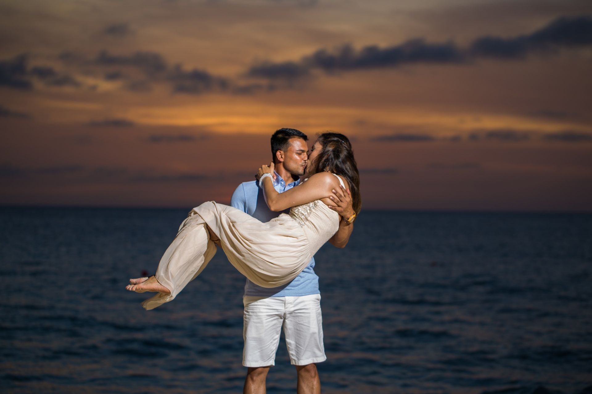 Couple embracing on a beach at sunset, with calm ocean and orange sky.