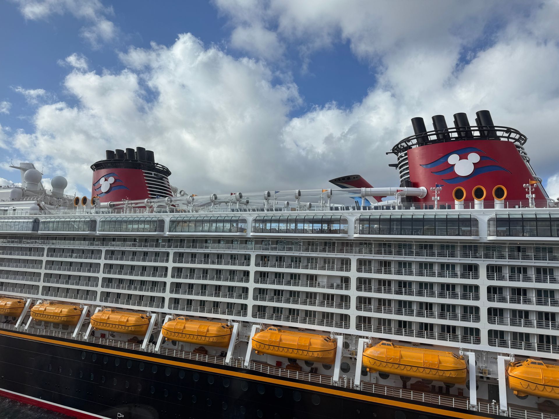 Disney cruise ship docked under cloudy sky, with red Mickey Mouse funnels and lifeboats along the side