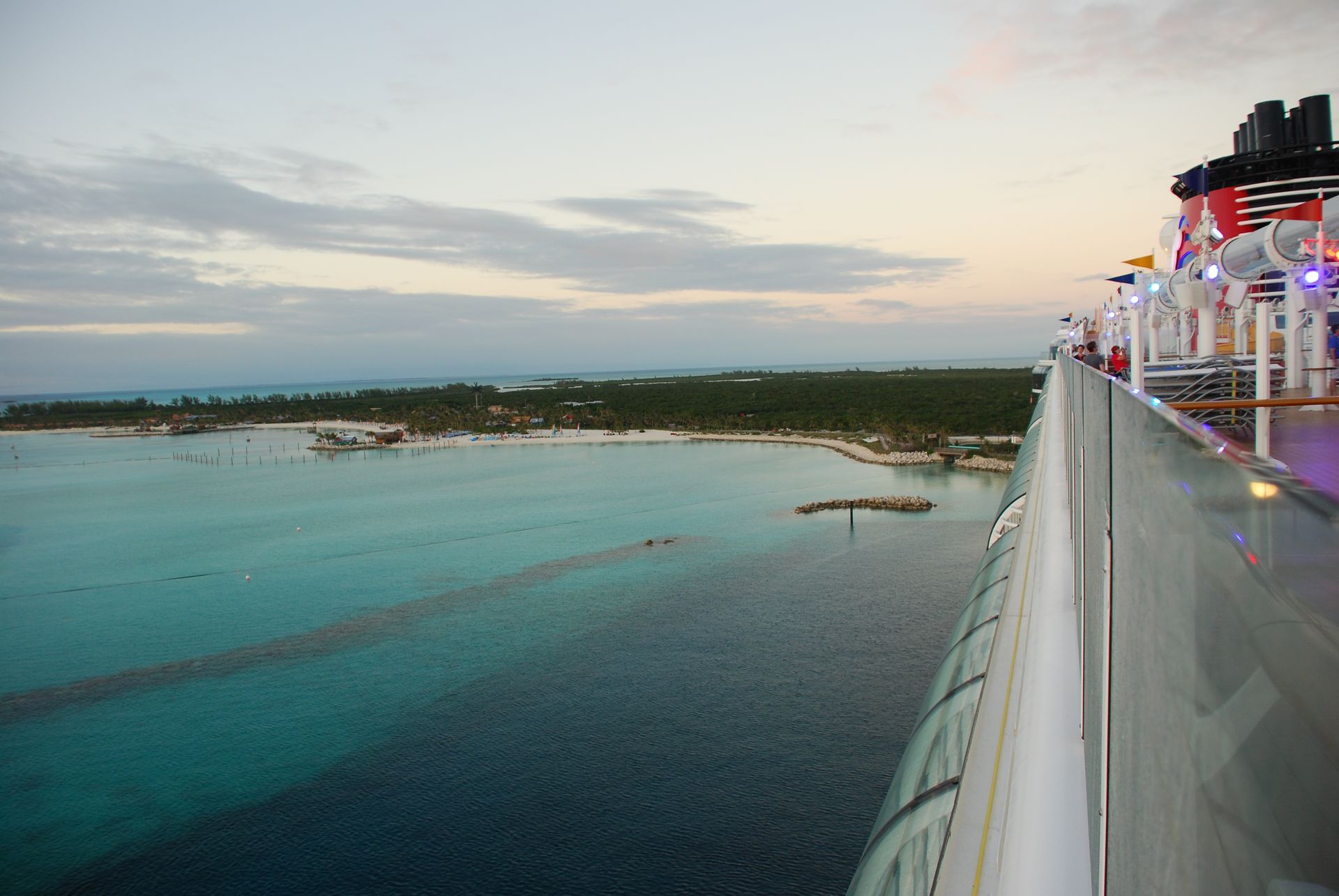 Cruise ship deck overlooking a turquoise bay at sunset, with distant shoreline and calm water