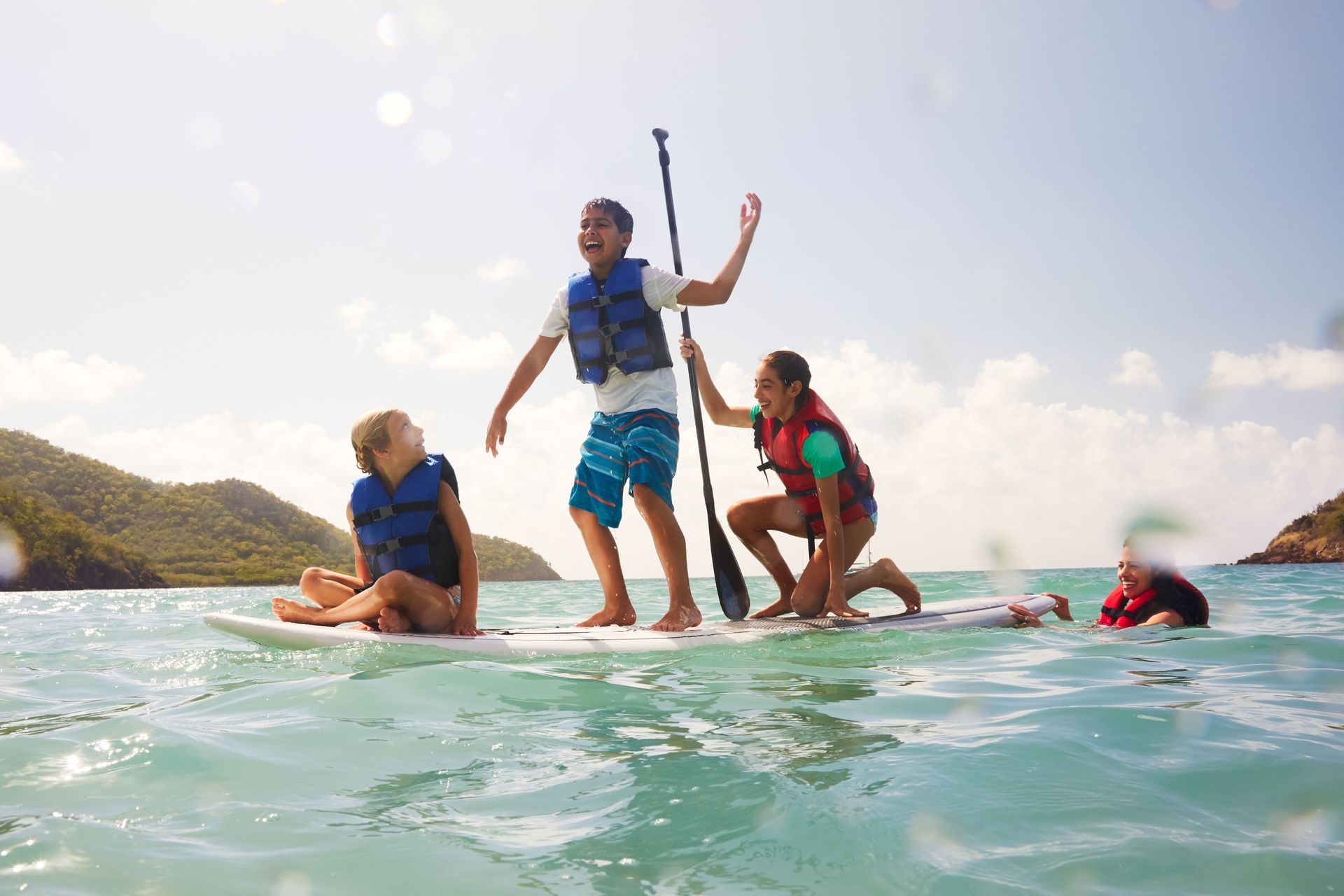 People standing and sitting on a paddleboard in turquoise water, with one swimmer nearby under a bright sky