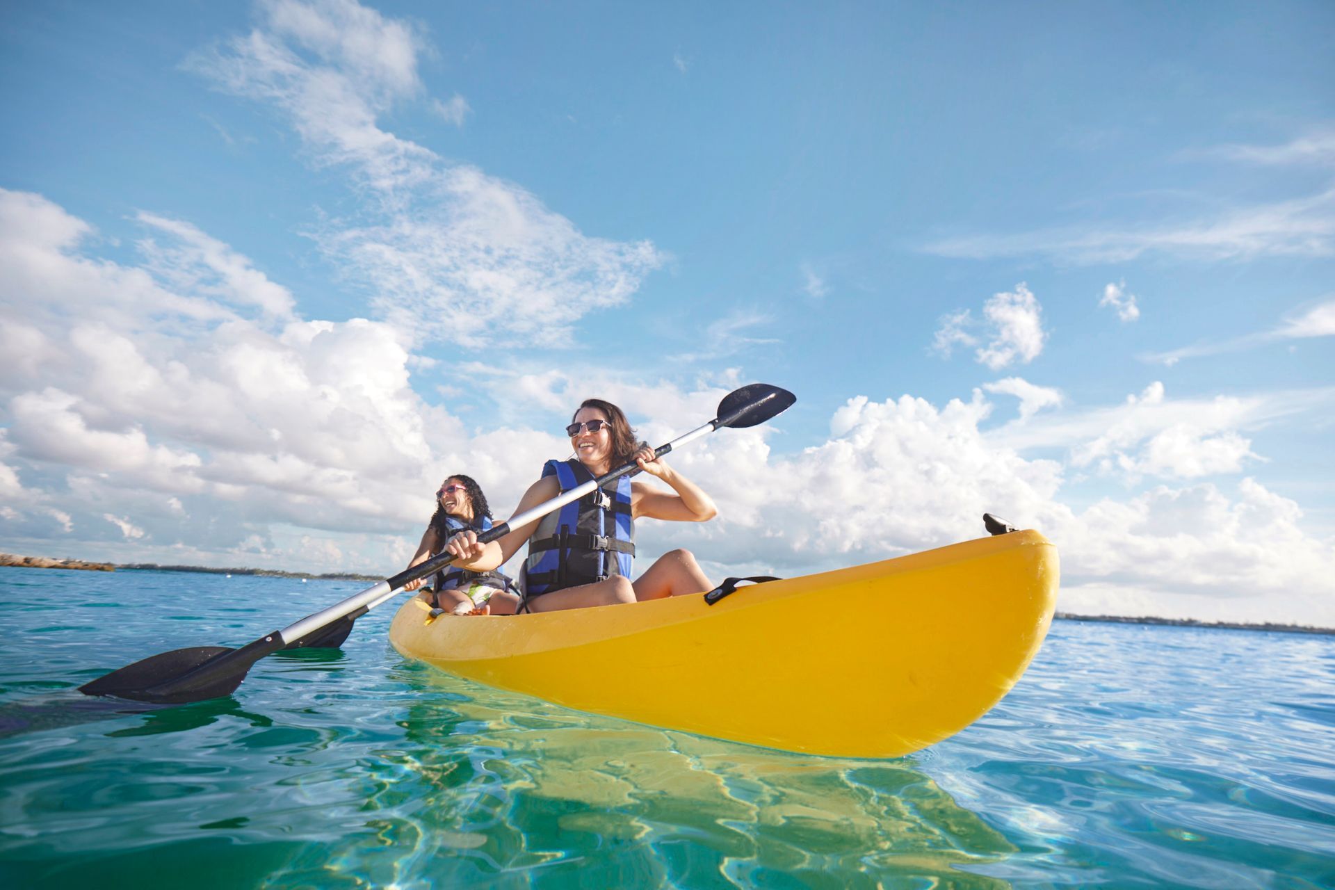Two people kayaking in a yellow kayak on turquoise ocean under a bright cloudy sky