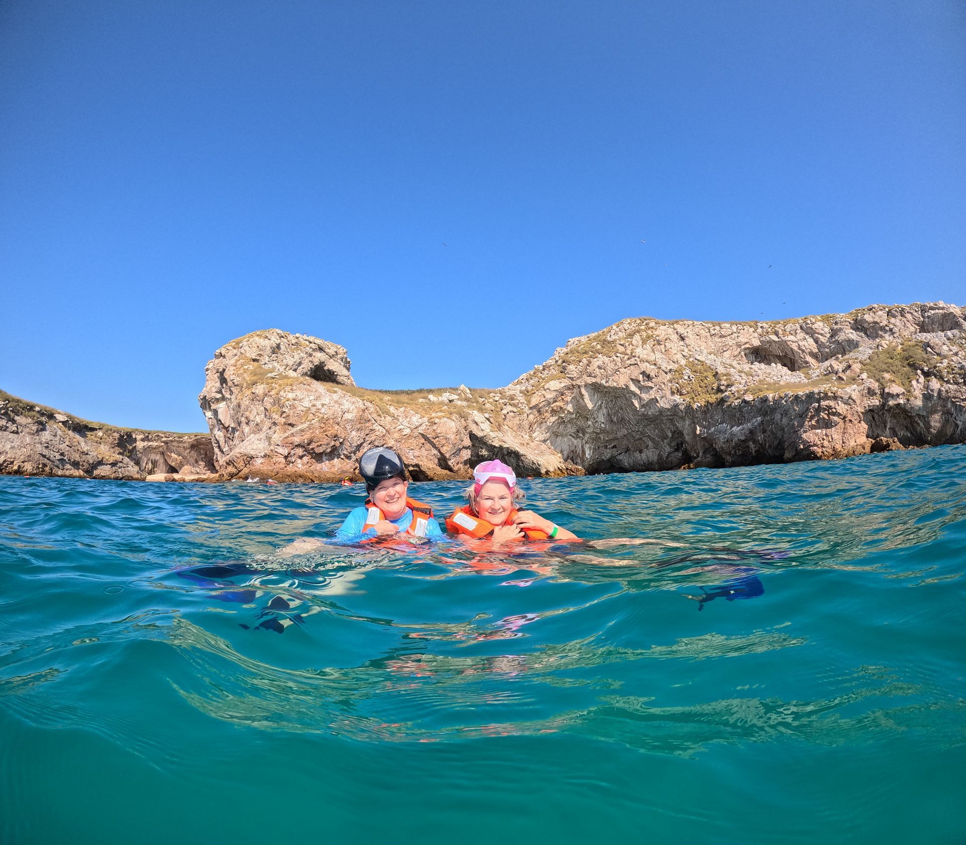 Two swimmers bob in turquoise water near rocky cliffs under a blue sky.