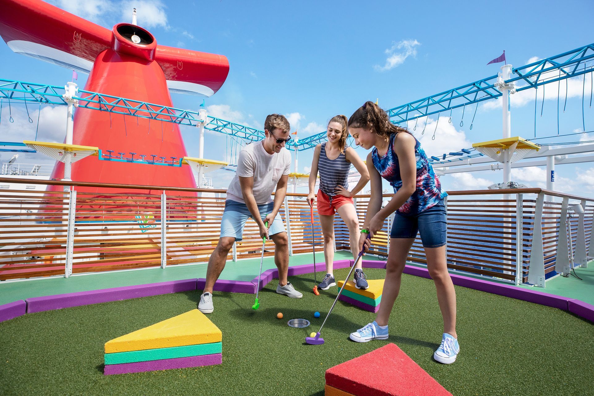 Three people playing mini-golf on a colorful cruise ship deck under a blue sky