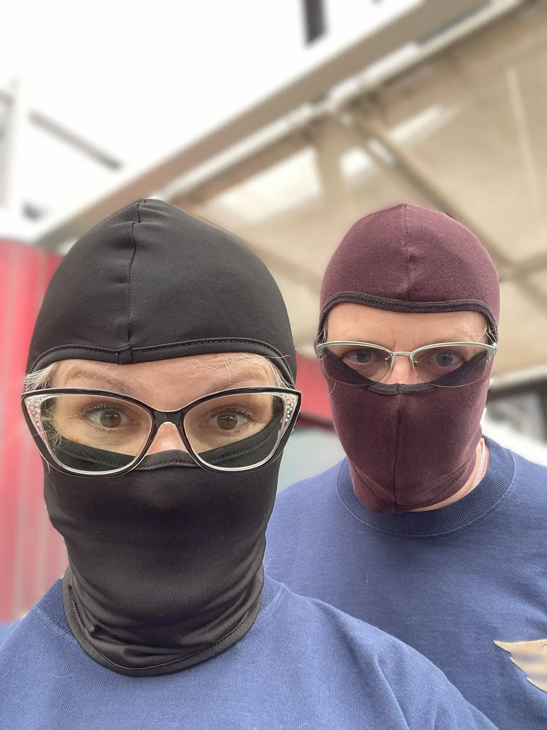 Two people in knit balaclavas and glasses, wearing blue shirts, indoors under bright lighting.