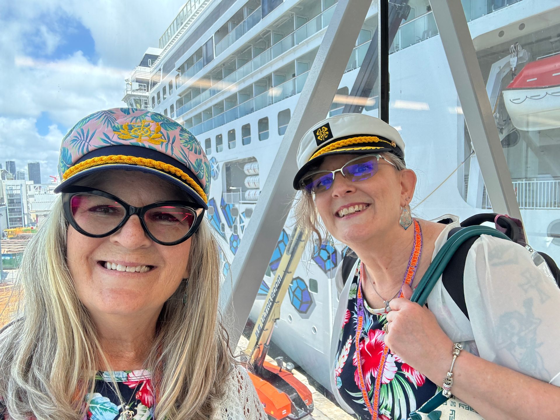 Two smiling people in colorful hats posing on a cruise ship deck with a city skyline behind them
