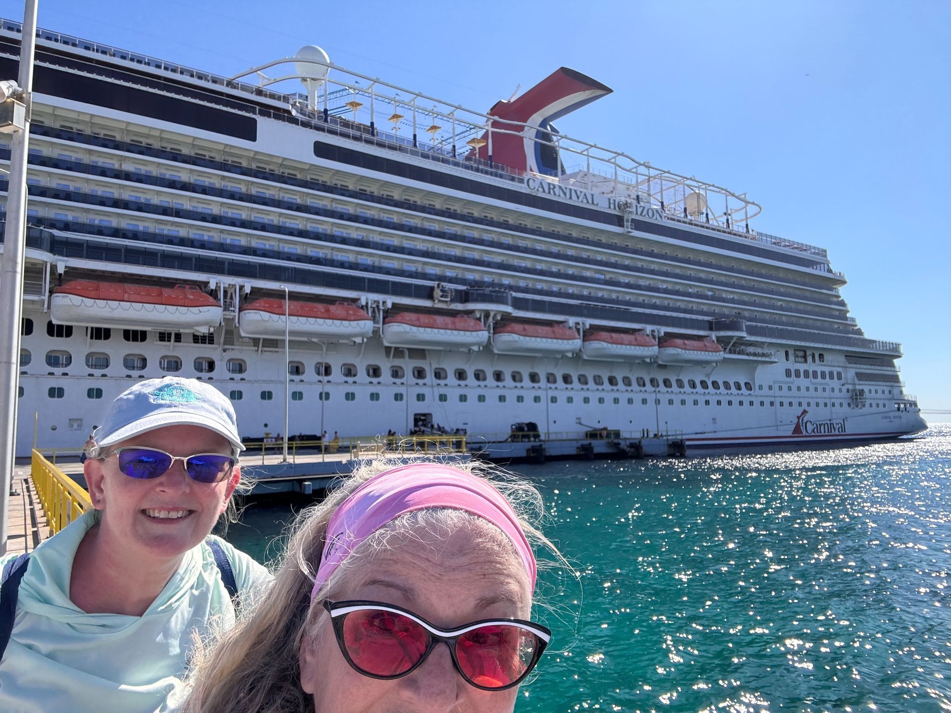 Two smiling people in sunglasses in front of a large cruise ship on bright blue water