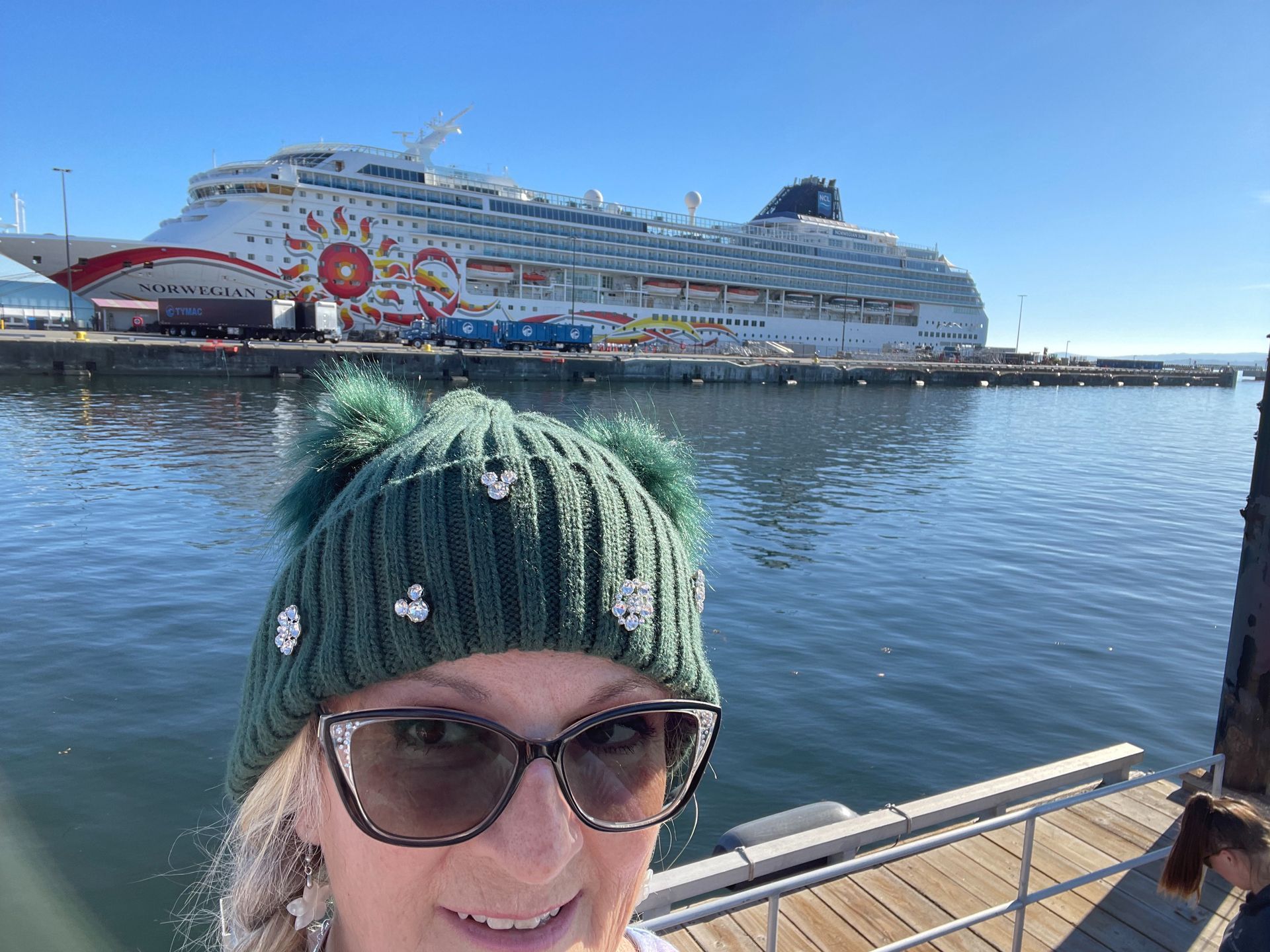 Selfie in a green knit hat and sunglasses with a cruise ship in the harbor behind.