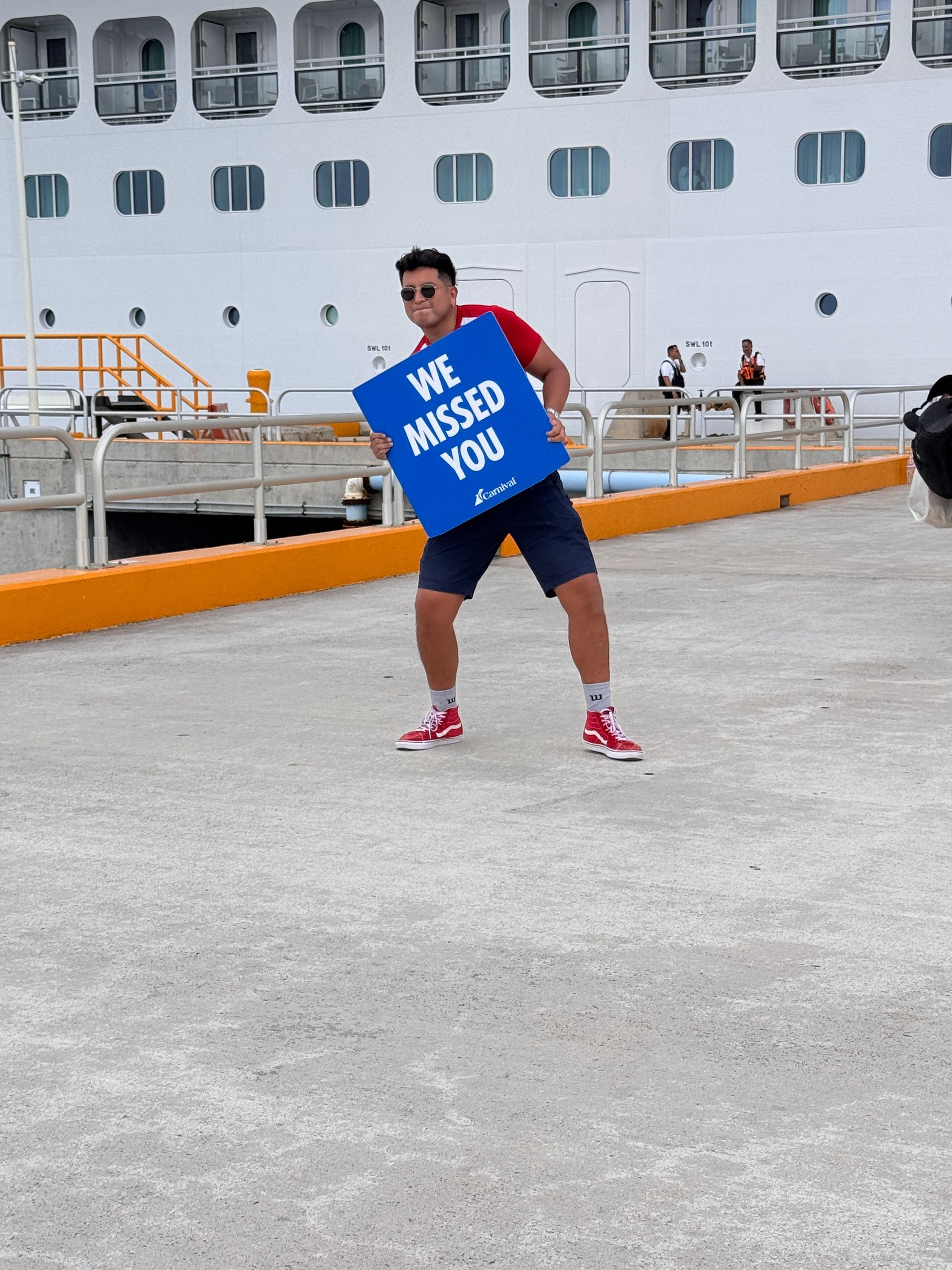 Person holding a blue “We Missed You” sign on a dock beside a large white cruise ship.