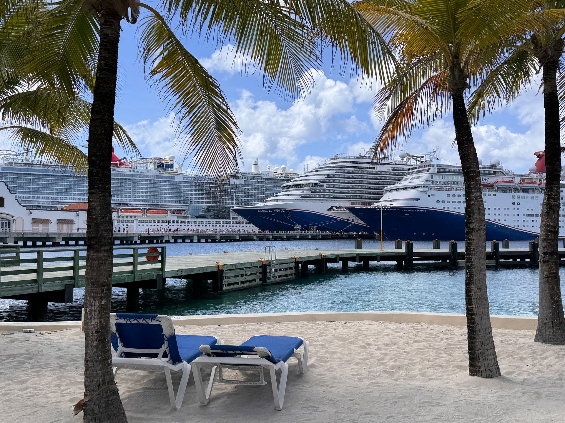 Cruise ship docked at a tropical beach with palm trees, lounge chairs, and blue water