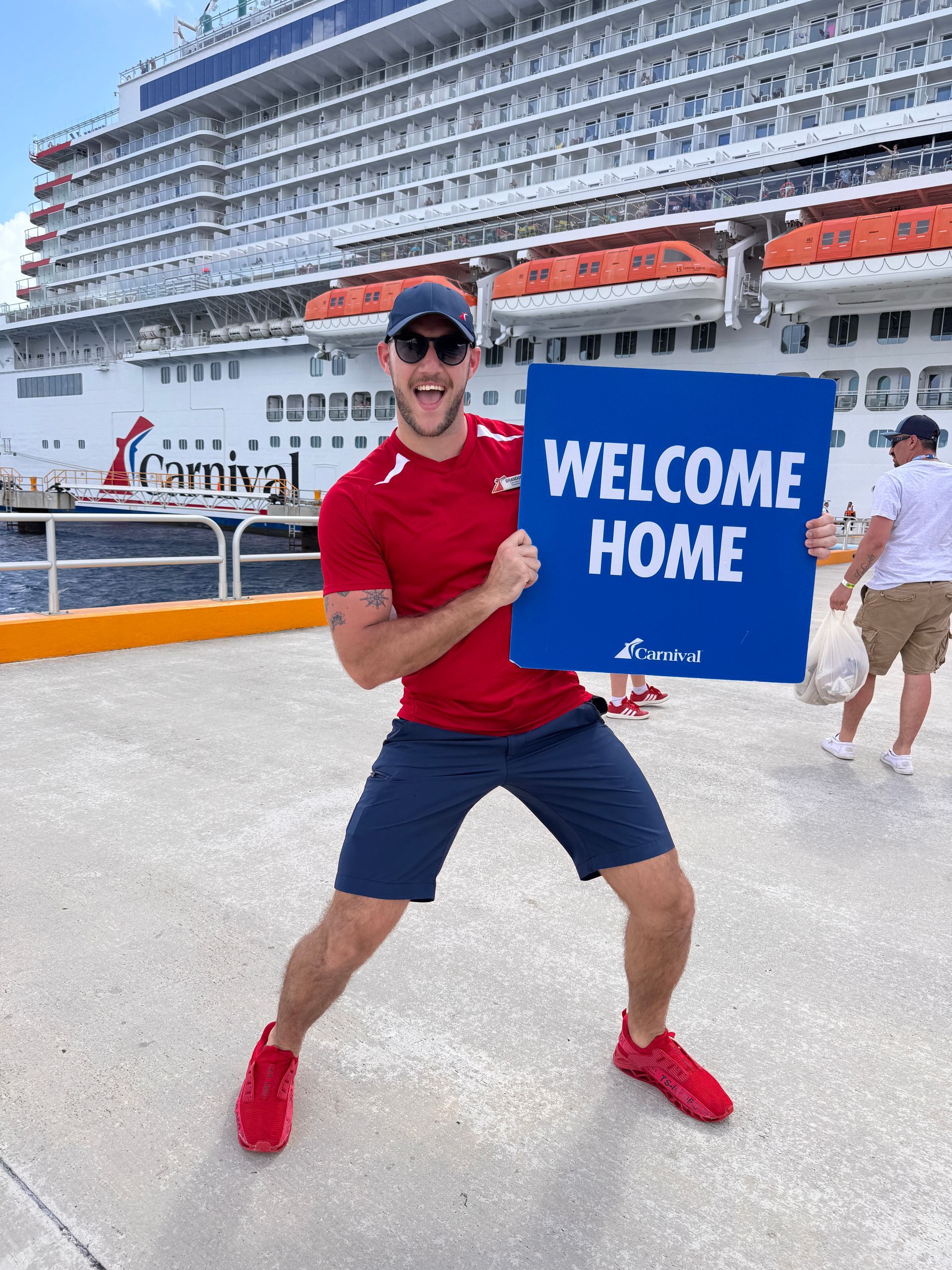 Cruise ship passenger in red shirt holding a blue “WELCOME HOME” sign on a sunny dock