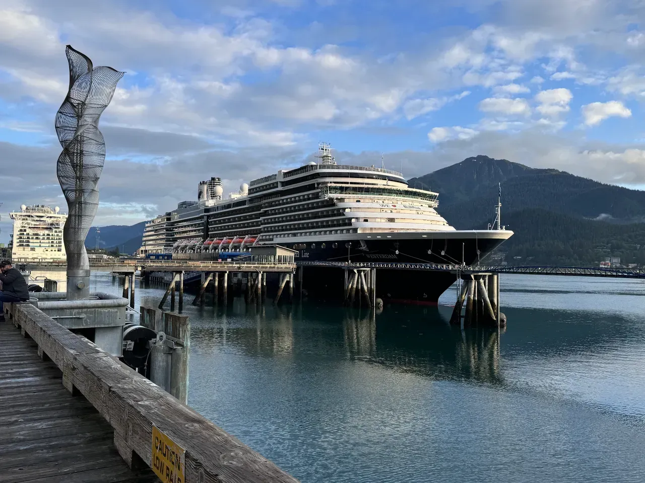 Cruise ship docked at a harbor beside a seaside promenade and sculpture, with mountains in the background.