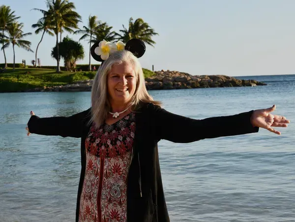 Smiling woman with arms outstretched on a beach, with palm trees and calm water behind her