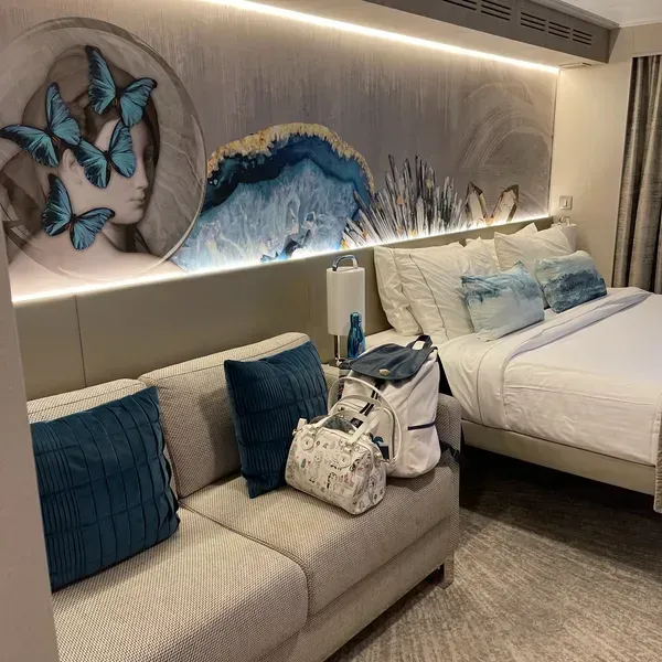 Cruise ship cabin with sofa, bed, blue butterfly-and-wave wall art, and luggage on the couch
