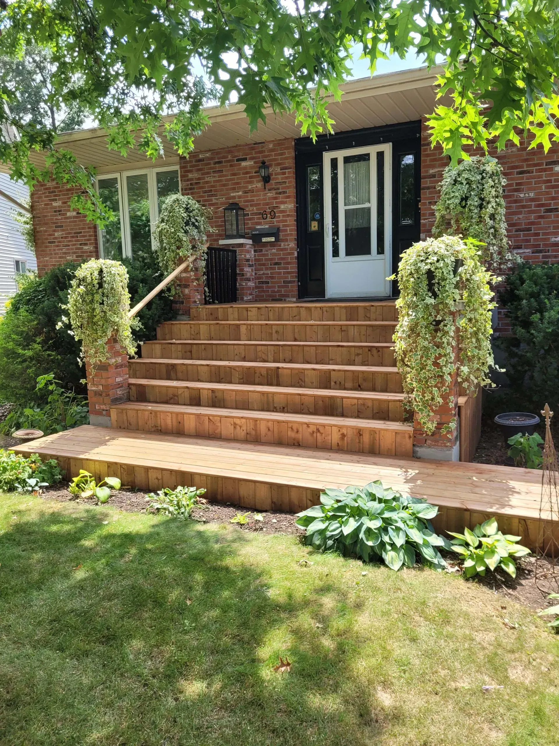 Brick house with wooden steps and hanging plants. Front yard with greenery and lawn.