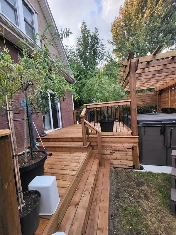Wooden backyard deck with steps, railing, and pergola beside a house and garden trees