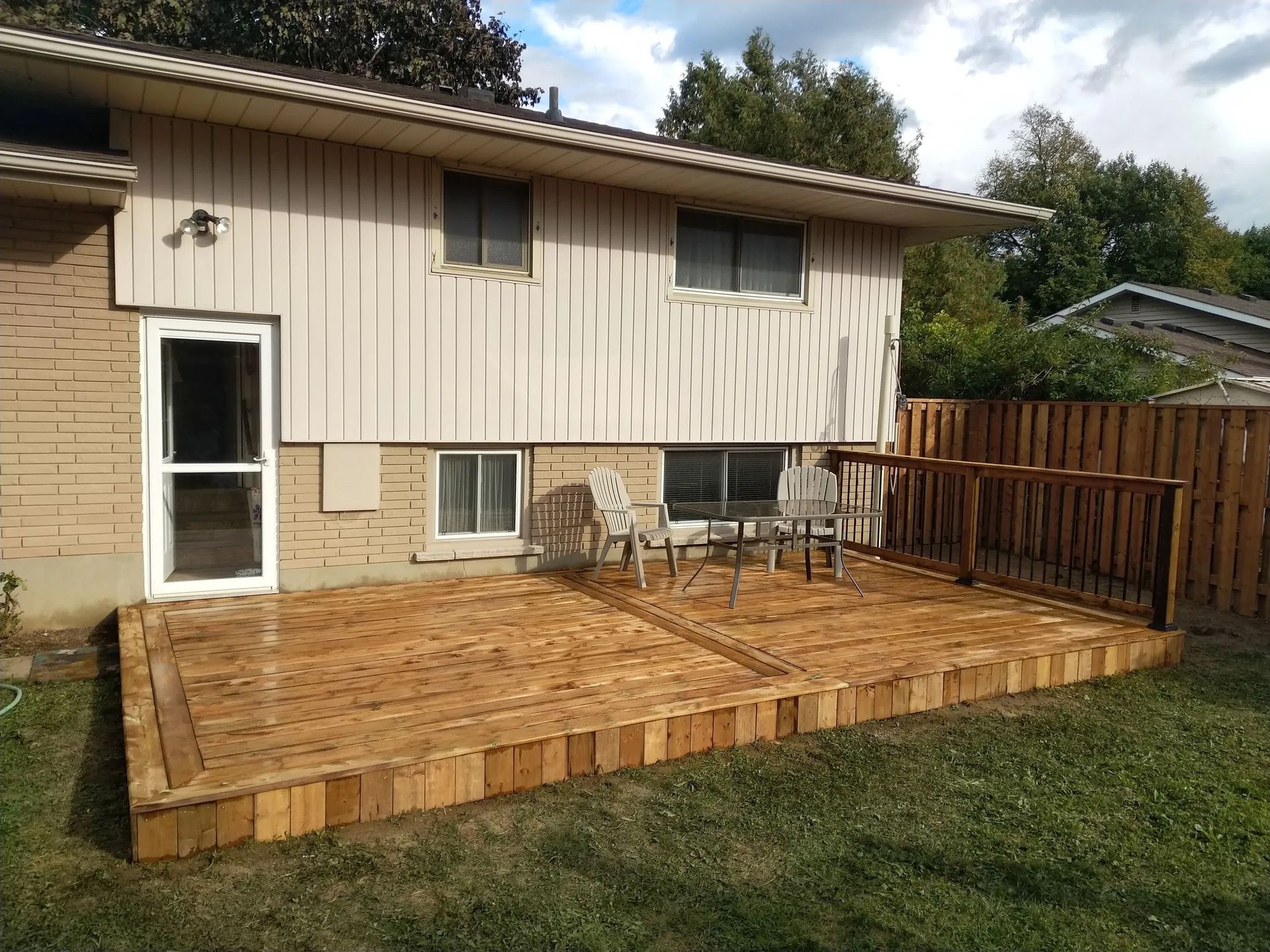 A wooden deck with furniture extends from a two-story house. A wooden fence surrounds the deck.