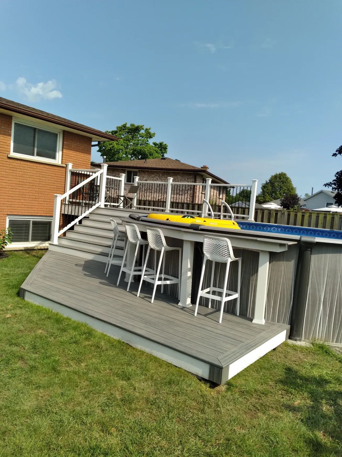 Backyard with above-ground pool, deck, and bar seating; grass lawn, brick house, blue sky.