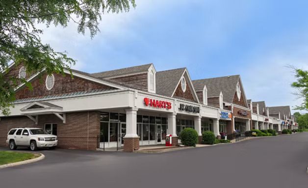 Shopping plaza exterior with storefronts, parked white SUV, and a red sign under a covered walkway