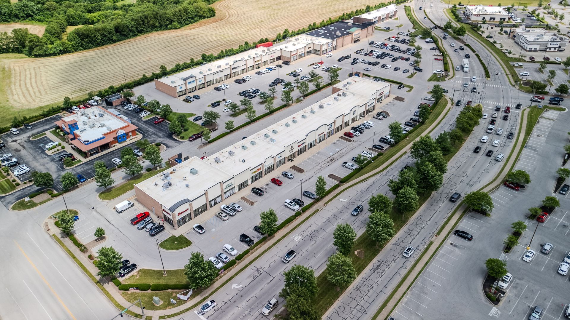 Aerial view of a shopping center with parking lots, roads, and cars surrounded by trees.