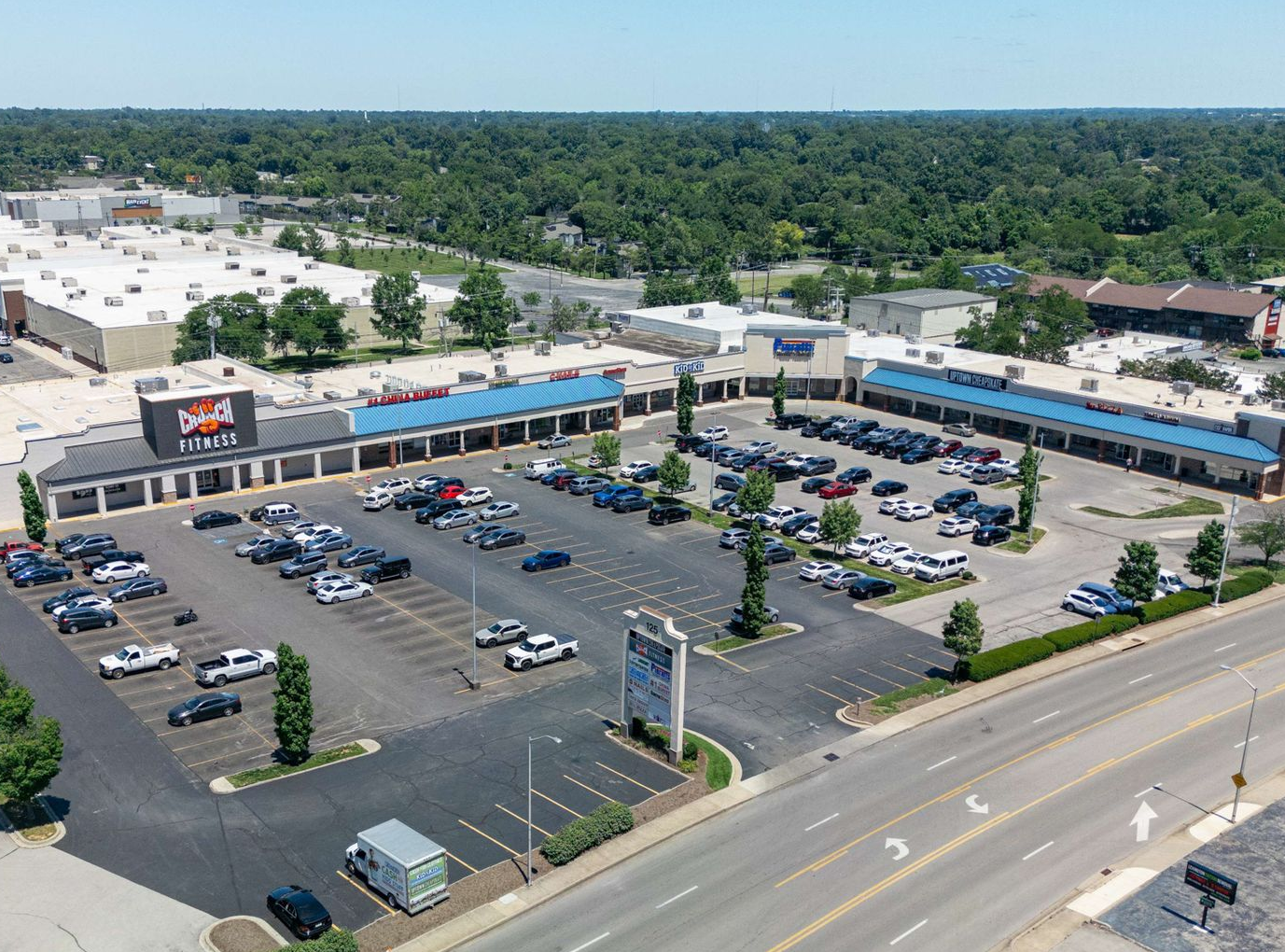 Aerial view of a busy shopping center with parking lots, storefronts, and surrounding trees