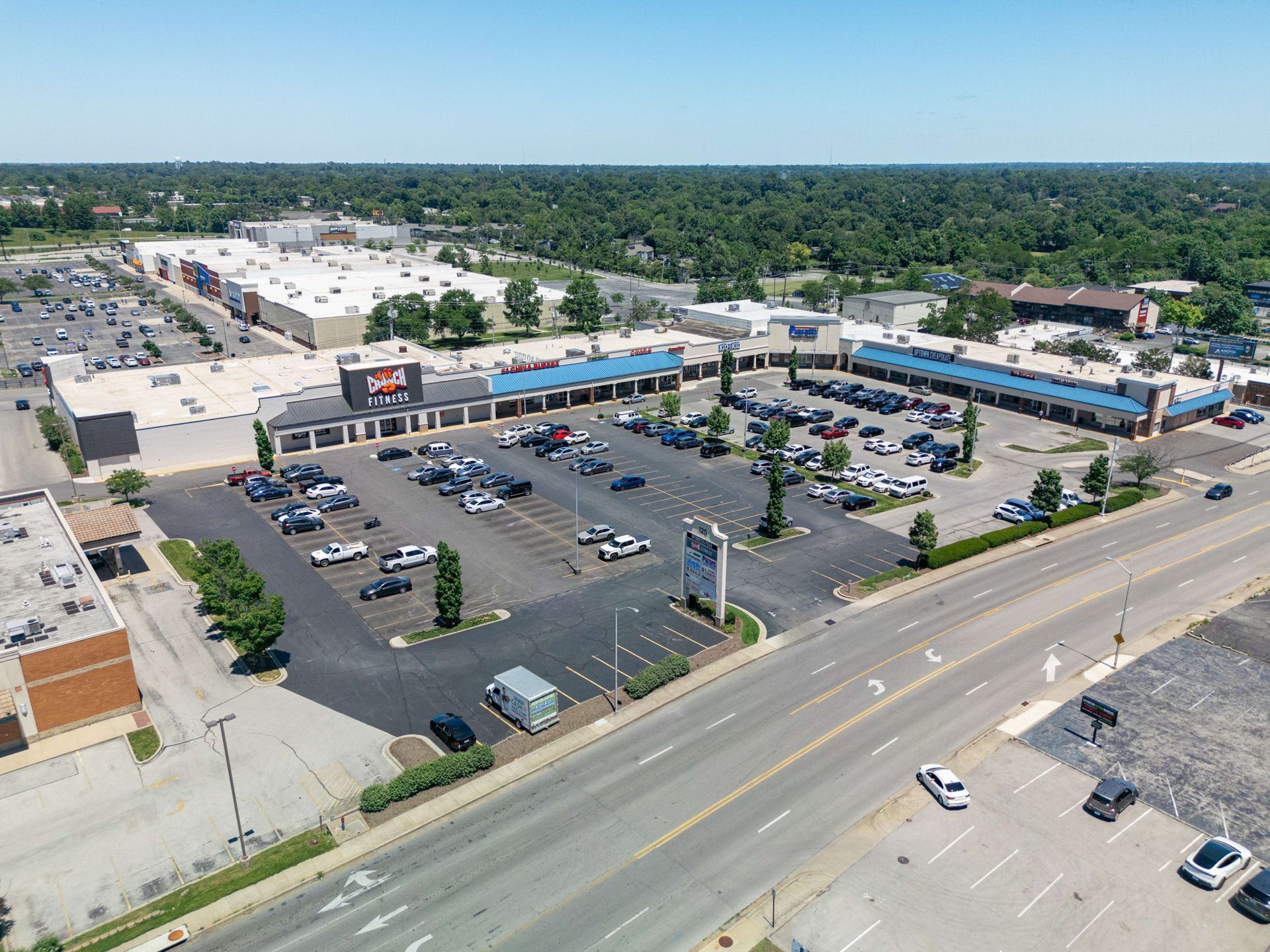 Aerial view of a shopping center with parking lot and surrounding trees along a roadside