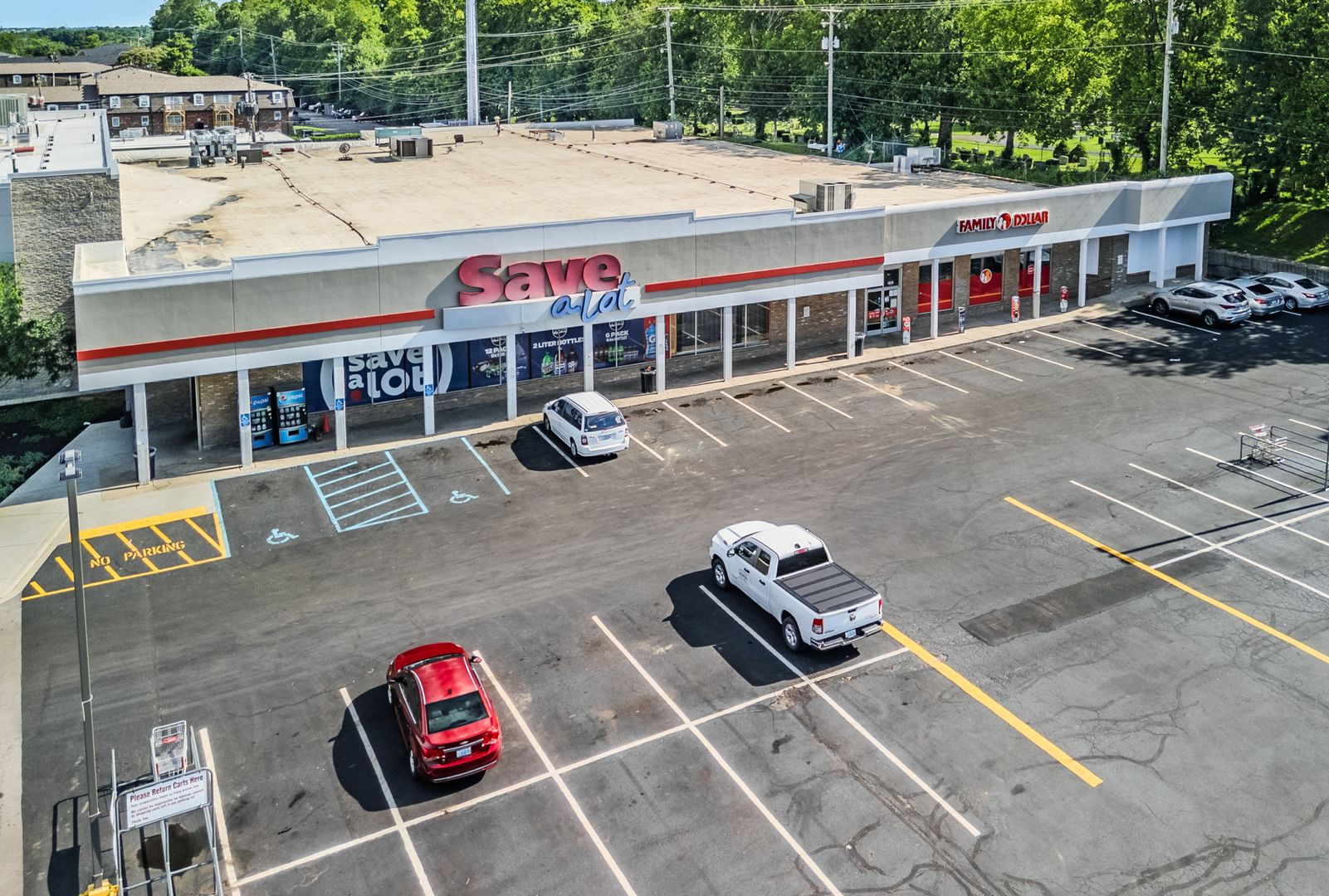 Shopping plaza exterior with storefronts, parked white SUV, and a red sign under a covered walkway
