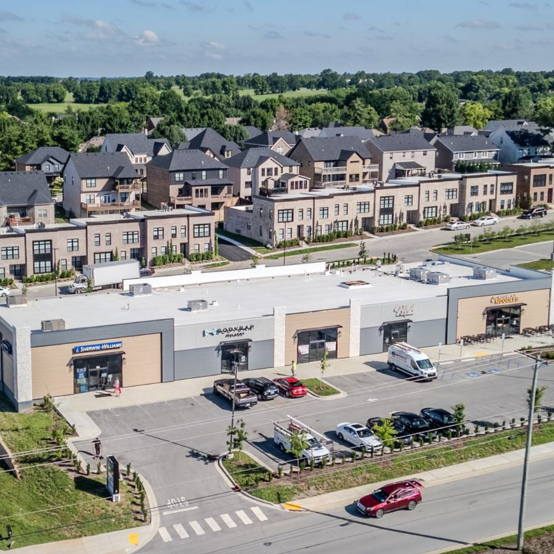 Aerial view of a suburban shopping center with parking lots, roads, and houses beyond