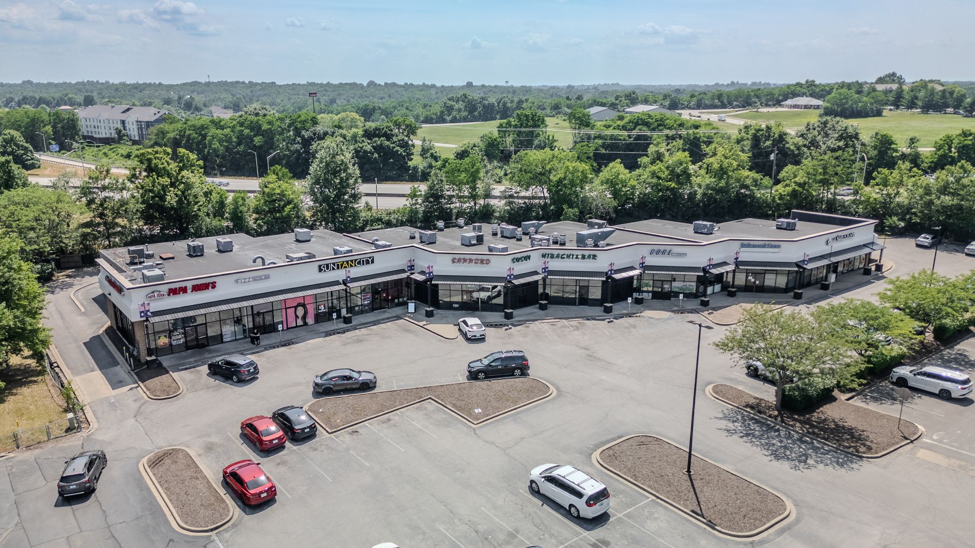 Aerial view of a strip mall with parked cars and surrounding trees under a clear sky