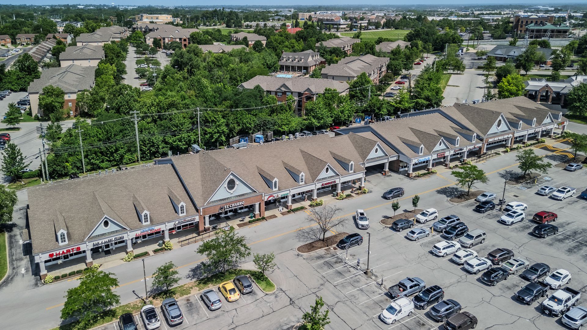 Aerial view of a shopping plaza with a parking lot, trees, and surrounding neighborhood homes.