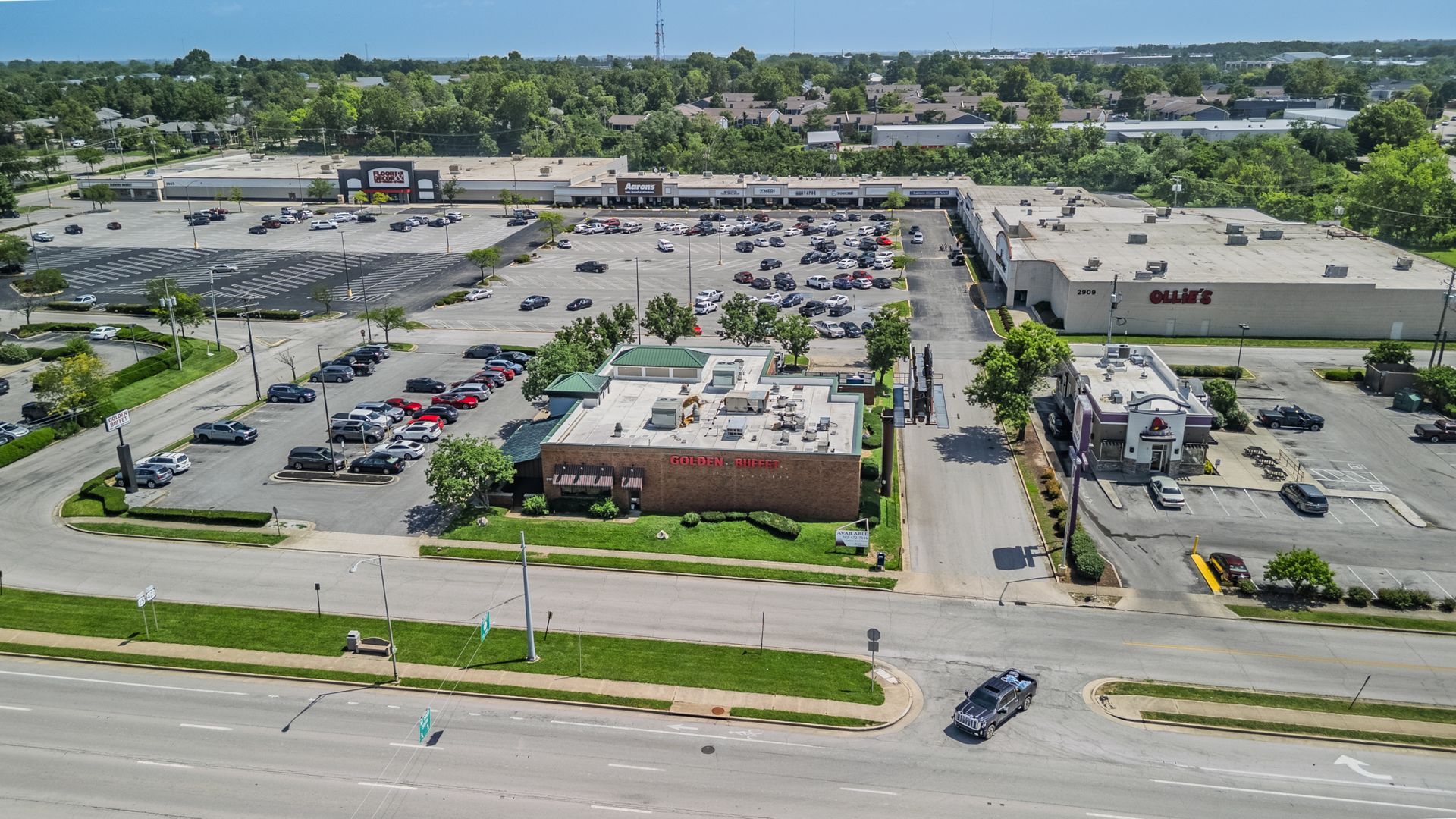 Aerial view of a shopping center parking lot with cars, storefronts, and a road in front.