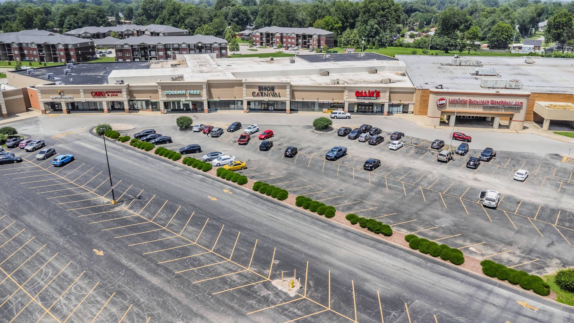 Aerial view of a busy shopping plaza with parked cars, surrounded by roads and trees.